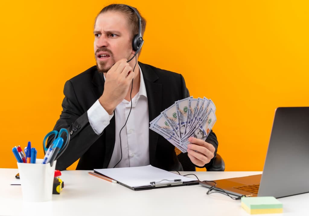 A man wearing a headset sits in front of a laptop and holds money in his hands