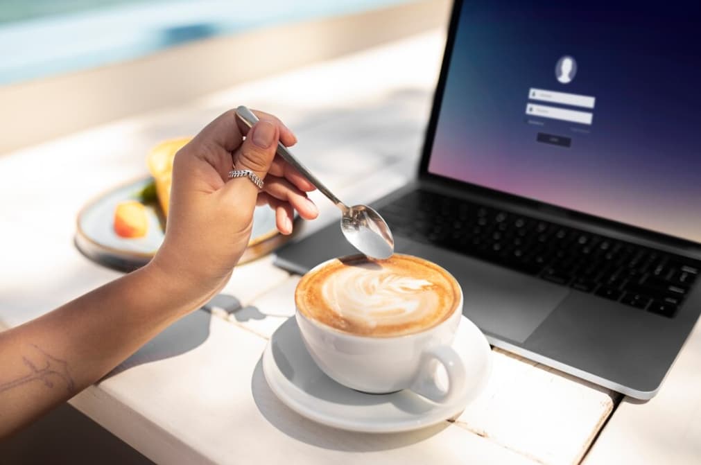 A person stirs a cup of cappuccino beside a laptop on a sunny table